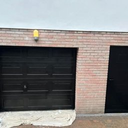 Damian Pietraszewicz - Pomarańczowy pojemnik i puszka farby obok czarnej bramy garażowej z ceglaną obudową i czarnych drzwi wejściowych, po malowaniu. Żółta lampa nad bramą.