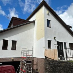 Damian Pietraszewicz - Malowanie elewacji domu jednorodzinnego w trakcie prac. Widoczny fragment rusztowania, częściowo pomalowana ściana i fragment samochodu. Słoneczny dzień.