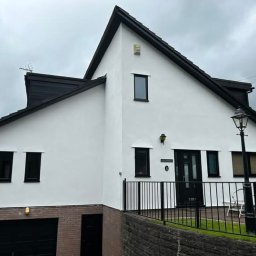 Damian Pietraszewicz - Elewacja domu po renowacji: białe ściany, czarna stolarka okienna i dachówka. Widoczny garaż z ceglaną podstawą i metalowe ogrodzenie.