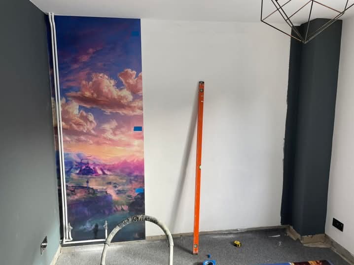 Wnętrze pokoju w trakcie prac: fototapeta z motywem krajobrazu częściowo naklejona na ścianę, obok poziomica i narzędzia, ściany w odcieniach szarości i bieli.