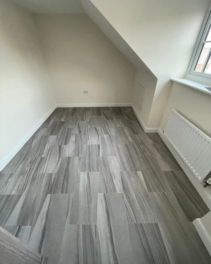 Pokój na poddaszu z nową, szarą podłogą imitującą kamień, świeżo po remoncie. Widoczne okno i grzejnik. Ukośne ściany dodają charakteru.