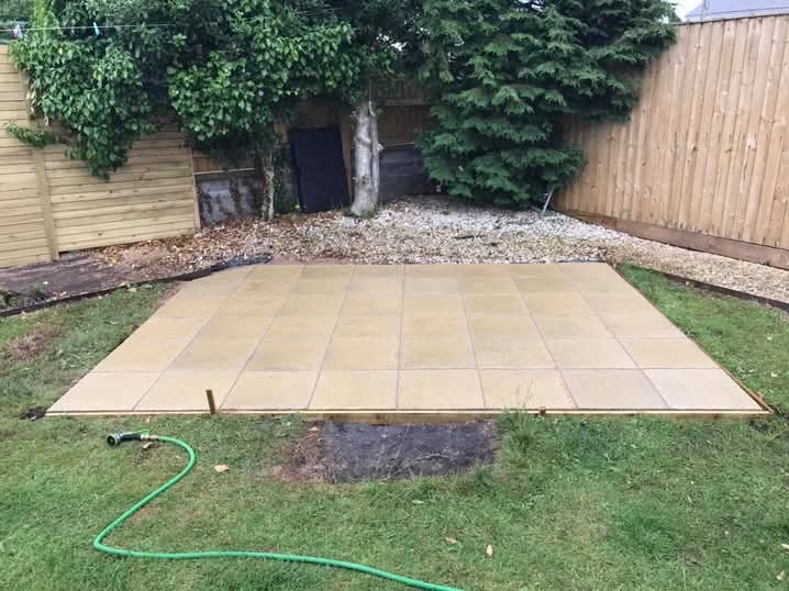 Nowo ułożony, jasny taras z płytek na trawniku, otoczony drewnianą ramą. W tle widoczne drzewa i drewniany płot. Wąż ogrodowy leży na trawie.