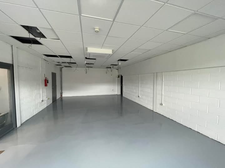 Wnętrze lokalu z szarą podłogą, białe ściany z bloków, sufit kasetonowy z ubytkami. Widoczny fragment drzwi i gaśnica. Oświetlenie sufitowe.