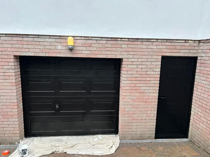 Pomarańczowy pojemnik i puszka farby obok czarnej bramy garażowej z ceglaną obudową i czarnych drzwi wejściowych, po malowaniu. Żółta lampa nad bramą.