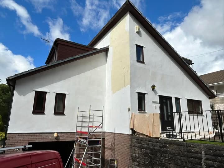 Malowanie elewacji domu jednorodzinnego w trakcie prac. Widoczny fragment rusztowania, częściowo pomalowana ściana i fragment samochodu. Słoneczny dzień.