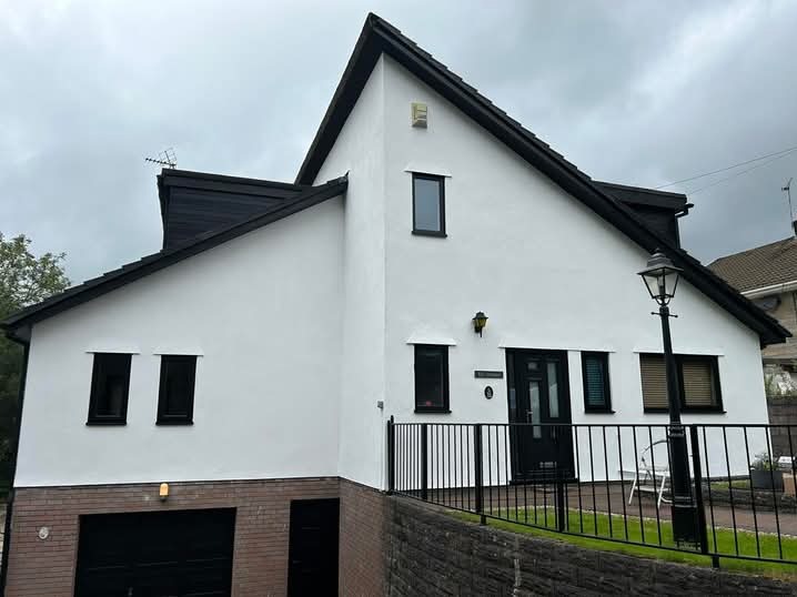 Elewacja domu po renowacji: białe ściany, czarna stolarka okienna i dachówka. Widoczny garaż z ceglaną podstawą i metalowe ogrodzenie.