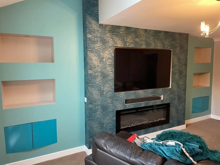 Ściana z wnękami i szafkami w odcieniach turkusu, ozdobna tapeta z motywem liści, telewizor i kominek elektryczny. Widok na salon po metamorfozie.