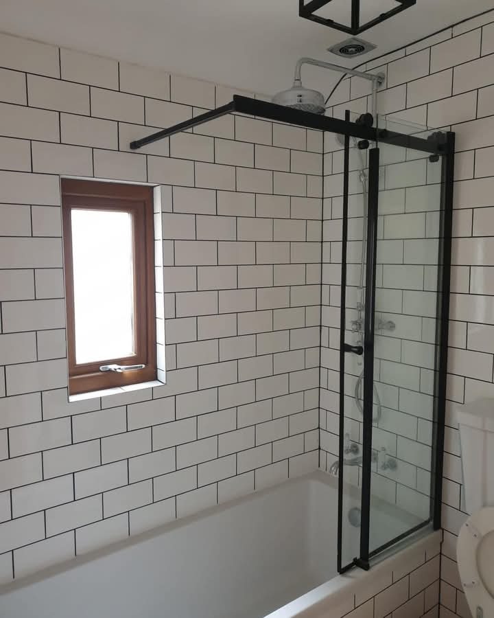 Nowoczesna łazienka z wanną i szklanym parawanem w czarnej ramie. Białe płytki imitujące cegłę z czarną fugą. Małe, drewniane okno. Minimalistyczny styl.