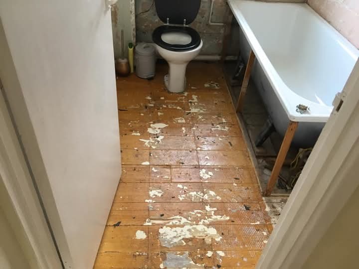 Łazienka w trakcie remontu: stara, drewniana podłoga z resztkami farby, toaleta i wanna na drewnianych podporach. Widok zza futryny drzwi.