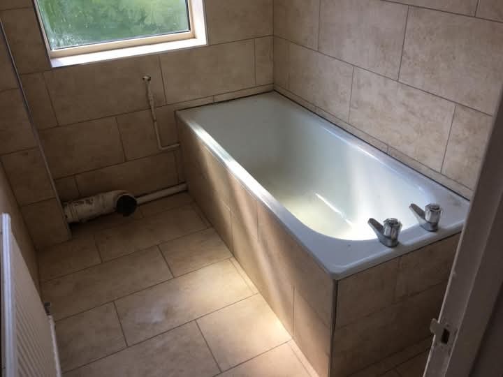 Wykończona łazienka z beżowymi płytkami: wanna obudowana płytkami, widoczne baterie, okno i fragment rury. Ukończona realizacja z dbałością o detale.