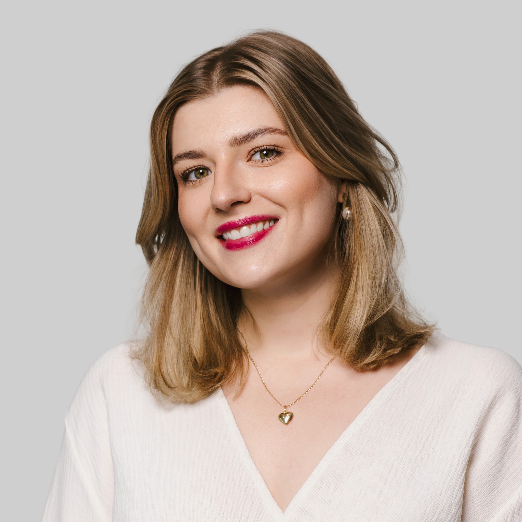 Portret uśmiechniętej blondynki z jaskrawą szminką, w białej bluzce i złotej biżuterii na jasnoszarym tle. Wyraziste rysy twarzy i naturalny makijaż.