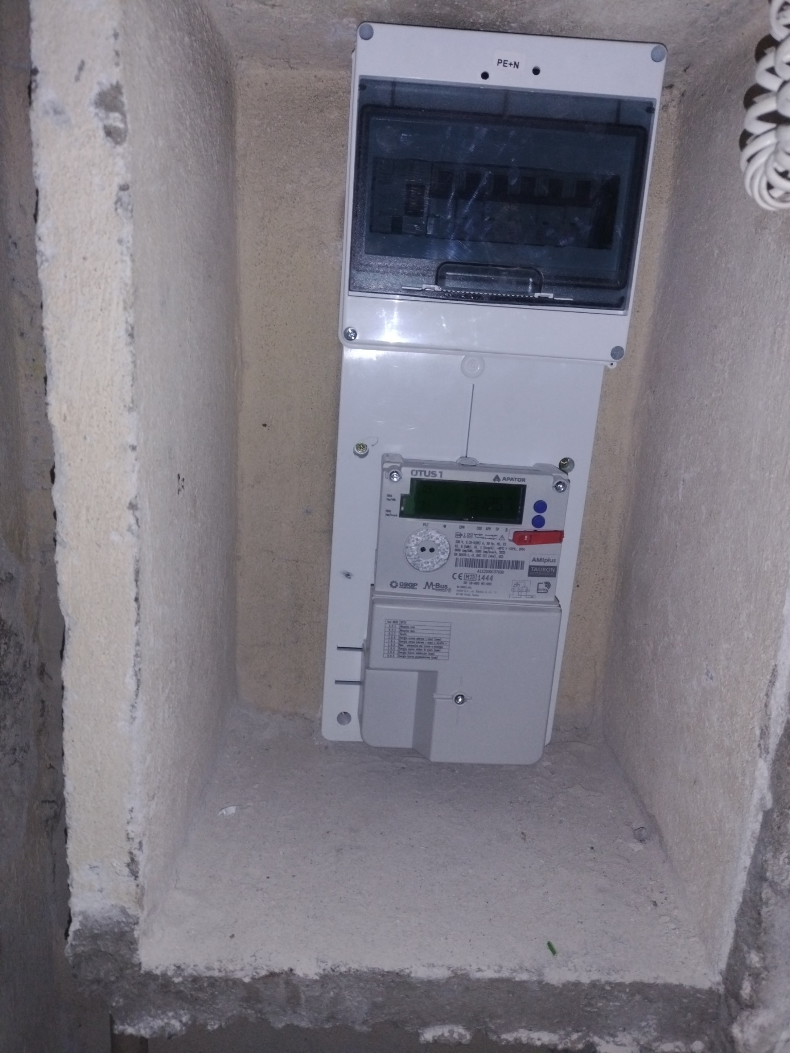 Zamontowana skrzynka z licznikiem elektrycznym w surowej wnęce. Widoczne ślady po kuciu w ścianie. Szara kolorystyka betonu i biała obudowa urządzeń.