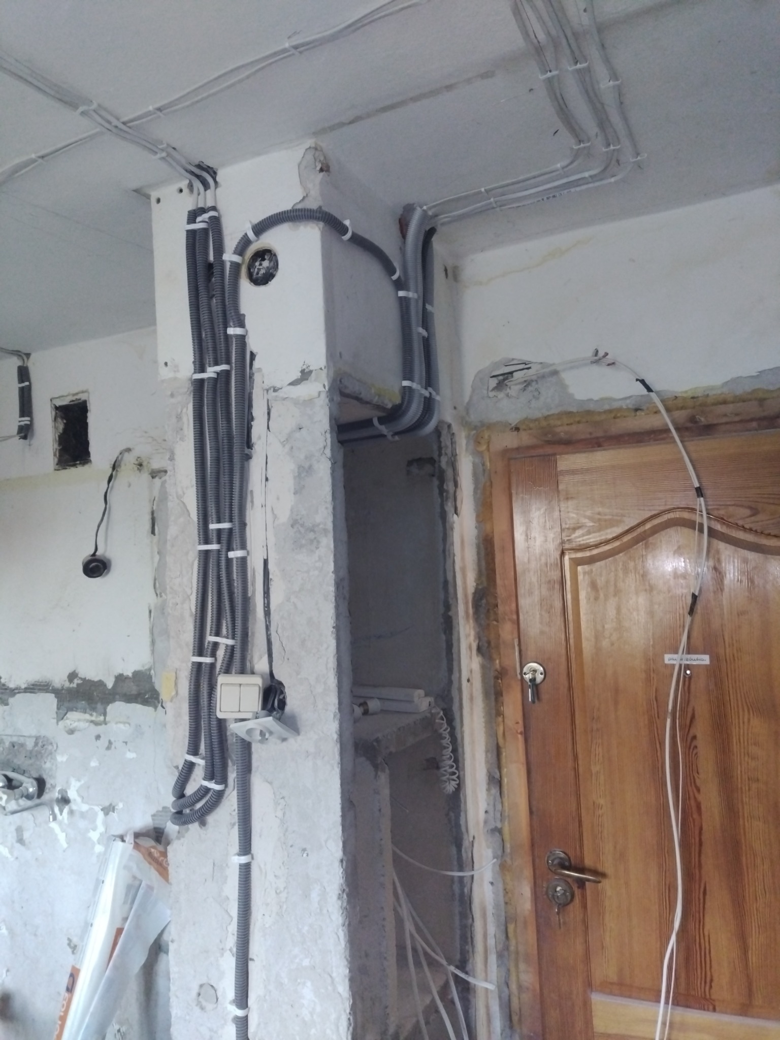 Szare rury z kablami przymocowane do ściany klamrami, widoczne gniazdka i włączniki. Ściana w trakcie remontu, obok drewniane drzwi z kluczem.