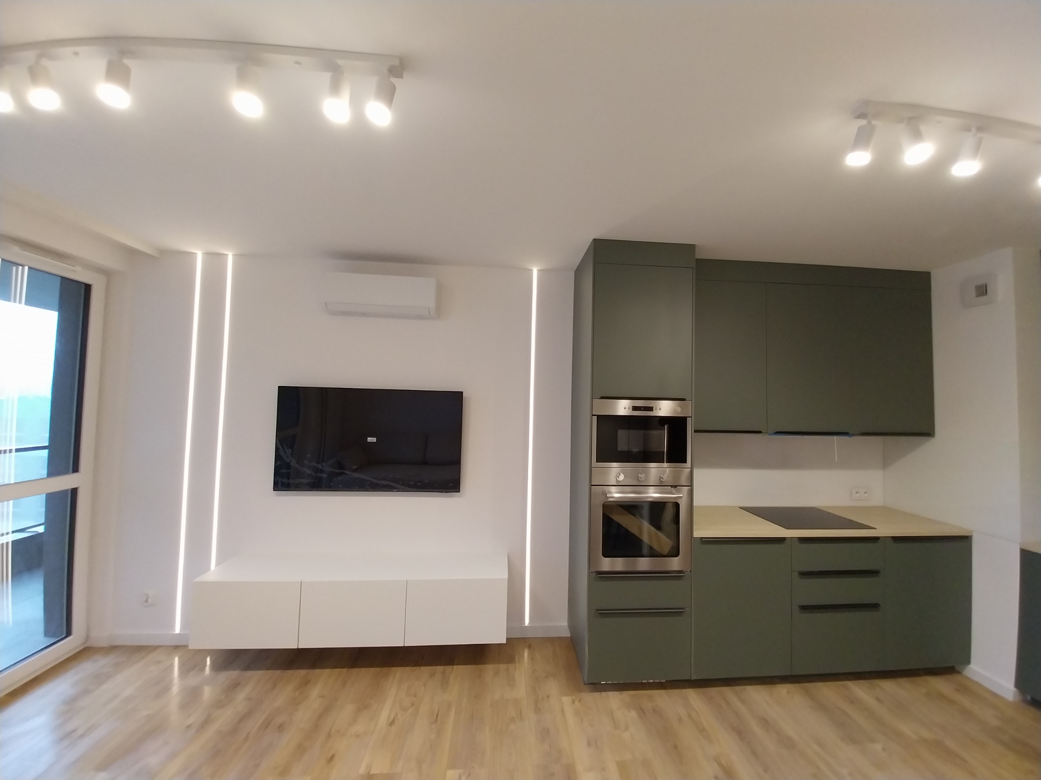 Nowoczesne wnętrze: drewniana podłoga, biała ściana z podświetleniem LED, szafka RTV, telewizor i oliwkowa zabudowa kuchenna z drewnianym blatem.