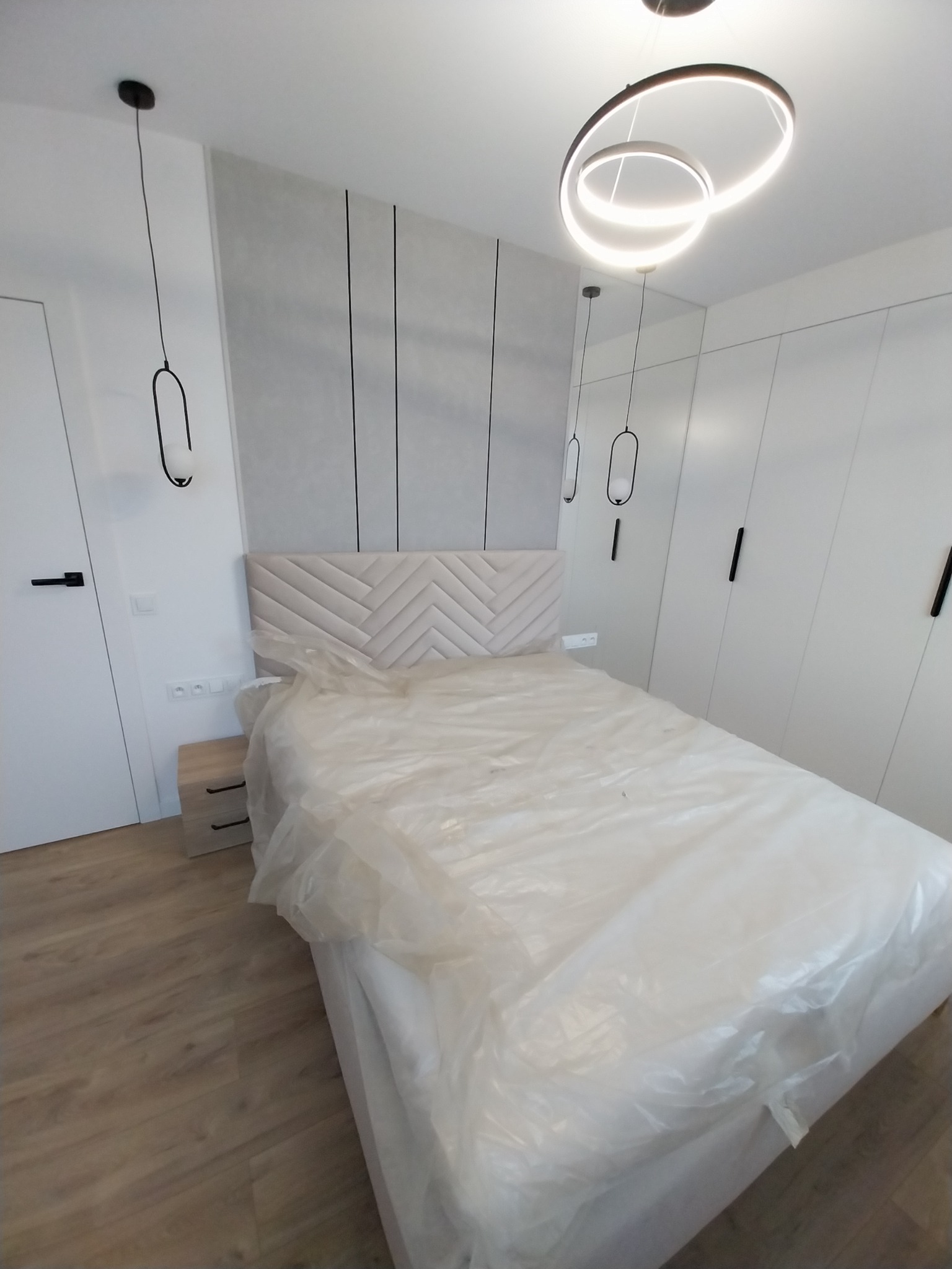 Nowoczesna sypialnia po remoncie: białe ściany z panelami, łóżko z zagłówkiem w folii ochronnej, designerskie lampy wiszące i szafa w zabudowie.