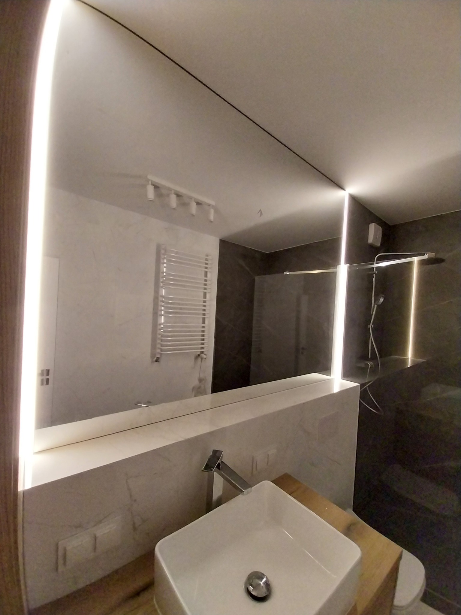 Nowoczesna łazienka z podświetlanym lustrem i marmurową umywalką. Widoczne detale armatury i oświetlenia punktowego, podkreślające minimalistyczny design wnętrza.