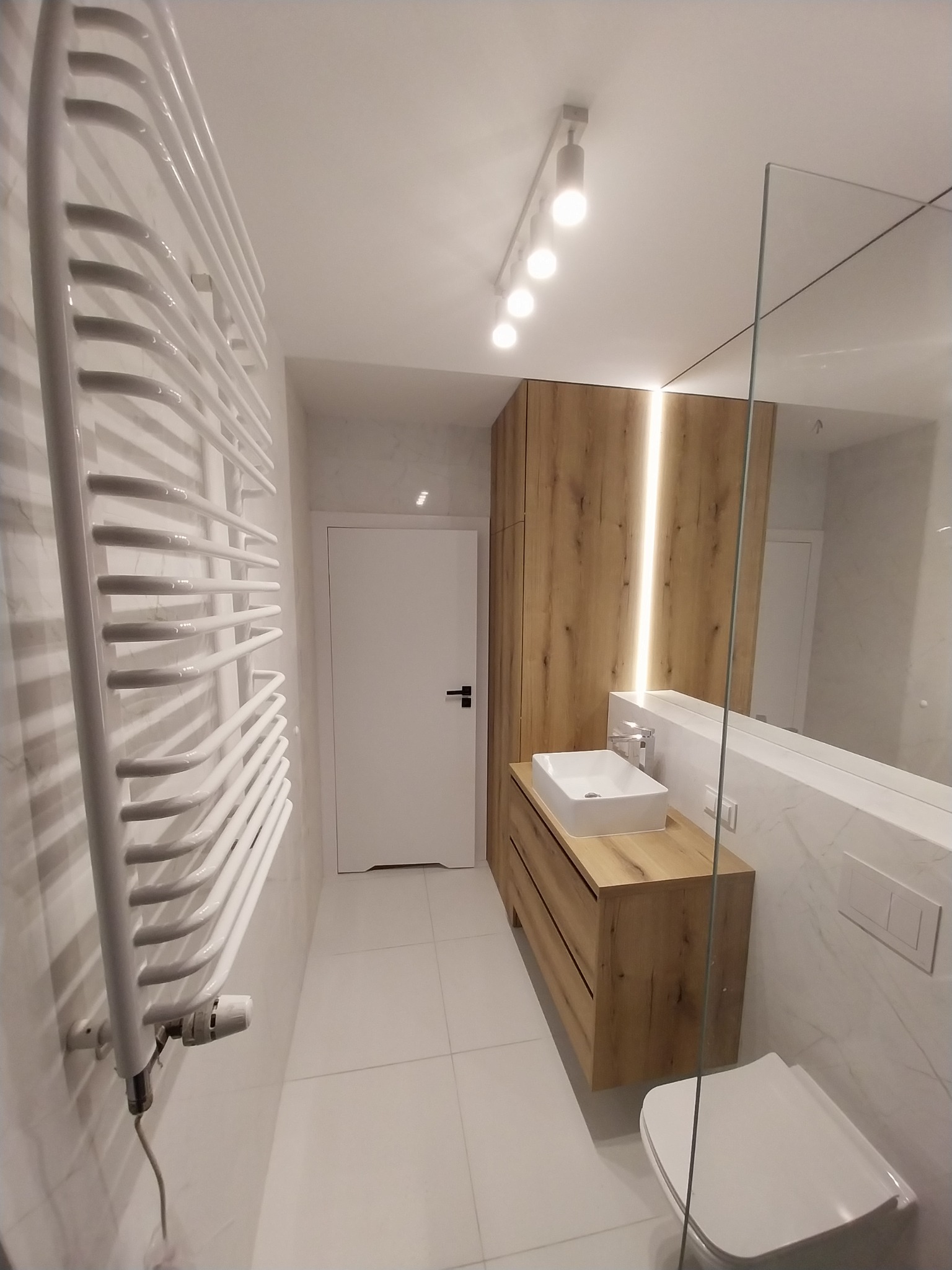 Nowoczesna łazienka z drewnianą szafką pod umywalką, białymi płytkami imitującymi marmur i grzejnikiem drabinkowym. Oświetlenie punktowe.