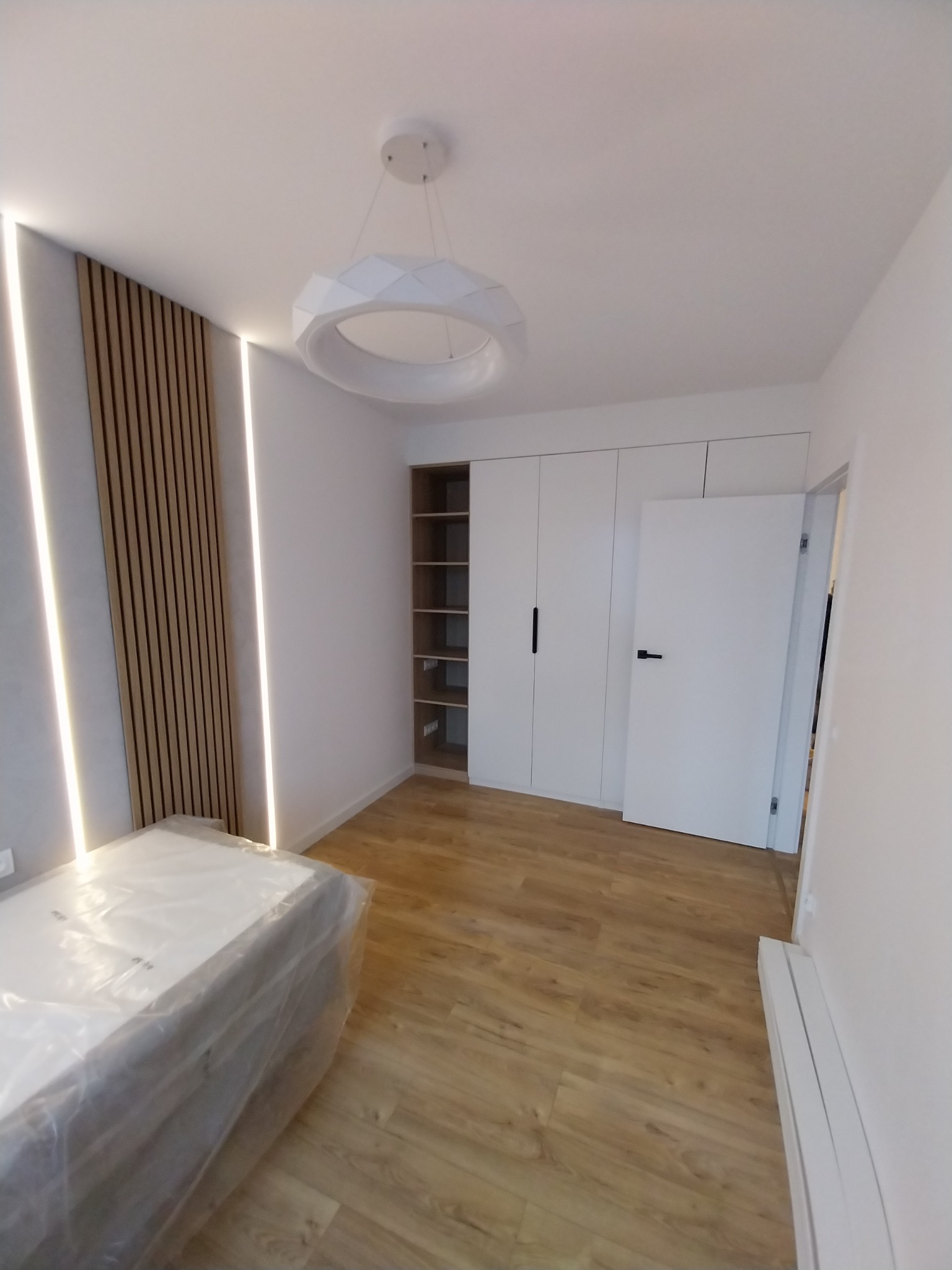 Jasne wnętrze pokoju po wykończeniu: drewniana podłoga, białe ściany, szafa wnękowa, dekoracyjny panel z lameli z oświetleniem LED i nowoczesna lampa sufitowa.