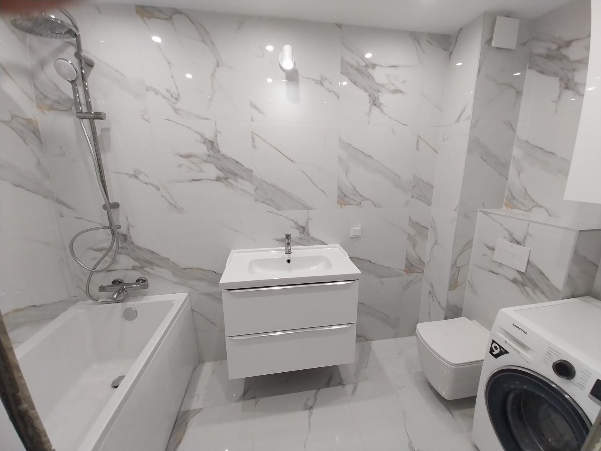 Nowoczesna łazienka z marmurowymi płytkami, białą armaturą i pralką. Minimalistyczny design, jasne oświetlenie, czyste linie. Umywalka z szafką, toaleta i wanna.