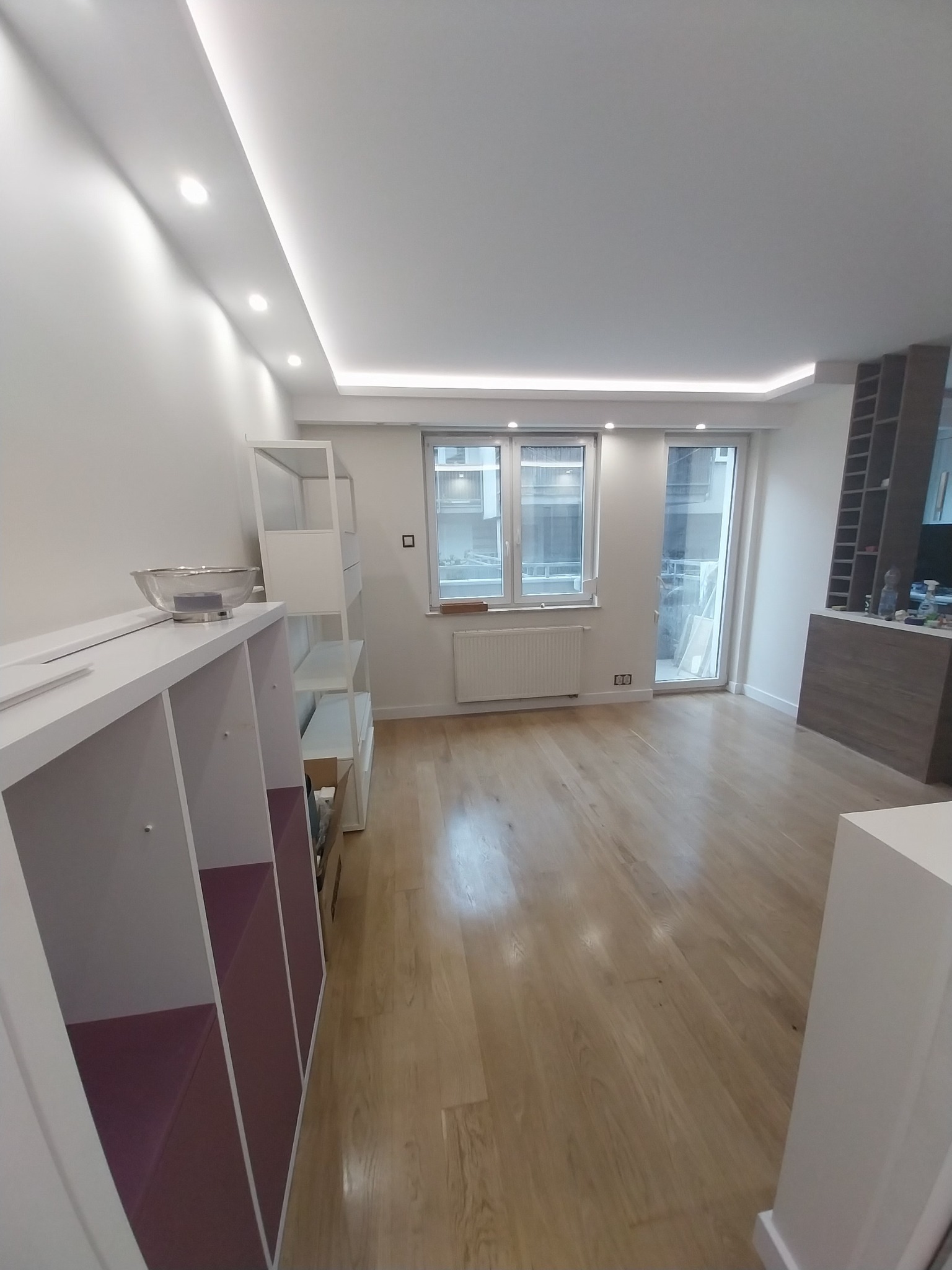 Jasne, przestronne wnętrze po remoncie: drewniana podłoga, białe ściany z oświetleniem LED, regały z fioletowymi akcentami, okno z widokiem na balkon.