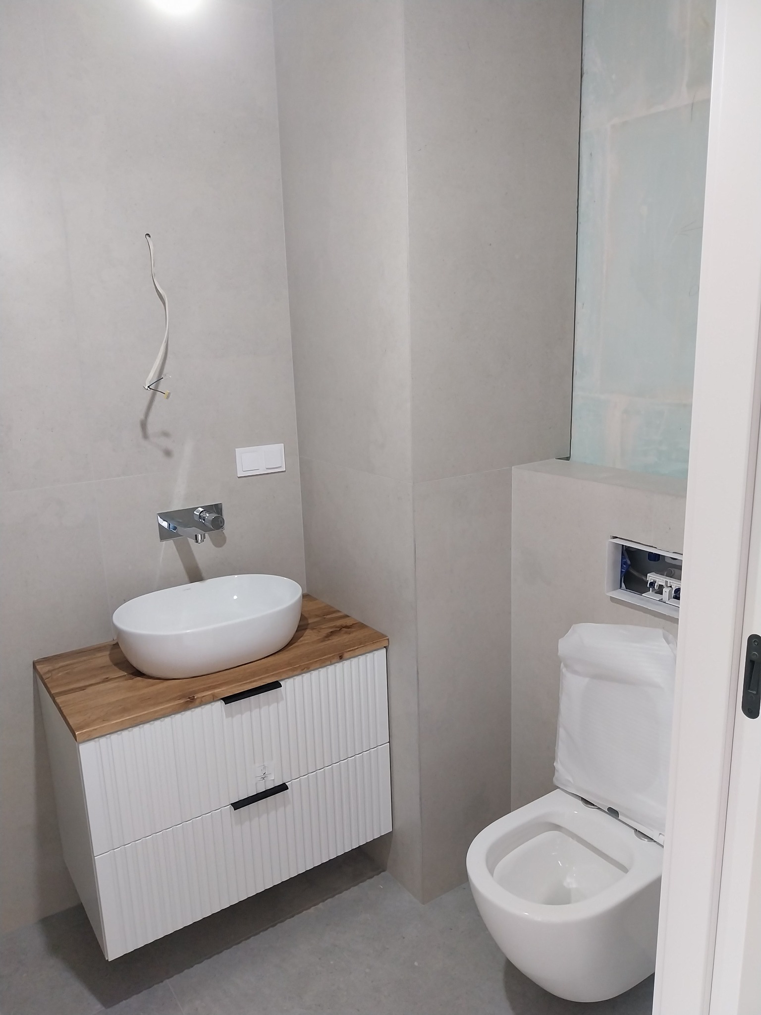 Nowoczesna łazienka z wiszącą toaletą i umywalką nablatową na szafce z drewnianym blatem. Minimalistyczny design, szare płytki, biała ceramika. Widoczne przewody.