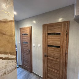 AZ DECOR - Dwa drewniane drzwi z przeszkleniami w szarej tonacji, na ścianie dekoracyjna faktura w odcieniach beżu i złota. Podłoga wyłożona szarymi panelami.