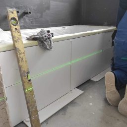 AZ DECOR - Układanie białych płytek na obudowie wanny, poziomica pionowa, linia lasera wyznacza poziom. Fragment osoby w niebieskim kombinezonie glazurnika.