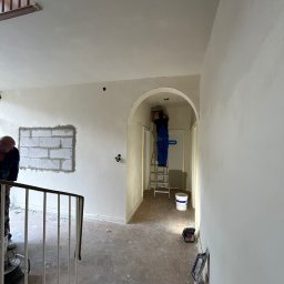 AZ DECOR - Wnętrze w trakcie prac wykończeniowych: szpachlowanie ścian przy użyciu drabiny, widoczny fragment ściany z bloków i narzędzia pracy. Jasne, naturalne światło.