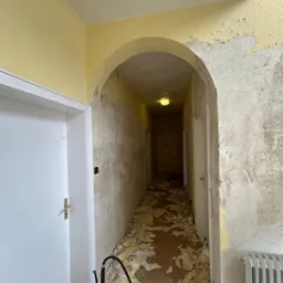 Korytarz w trakcie usuwania starej farby ze ścian i sufitu, widoczne resztki farby na podłodze, narzędzie do usuwania farby przy drzwiach, grzejnik i łukowe przejście.