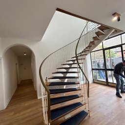 AZ DECOR - Spiralne schody z drewnianymi stopniami i białymi balustradami w jasnym wnętrzu. Widoczny fragment podłogi i okna z widokiem na zewnątrz. Osoba w tle.