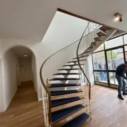 Spiralne schody z drewnianymi stopniami i białymi balustradami w jasnym wnętrzu. Widoczny fragment podłogi i okna z widokiem na zewnątrz. Osoba w tle.