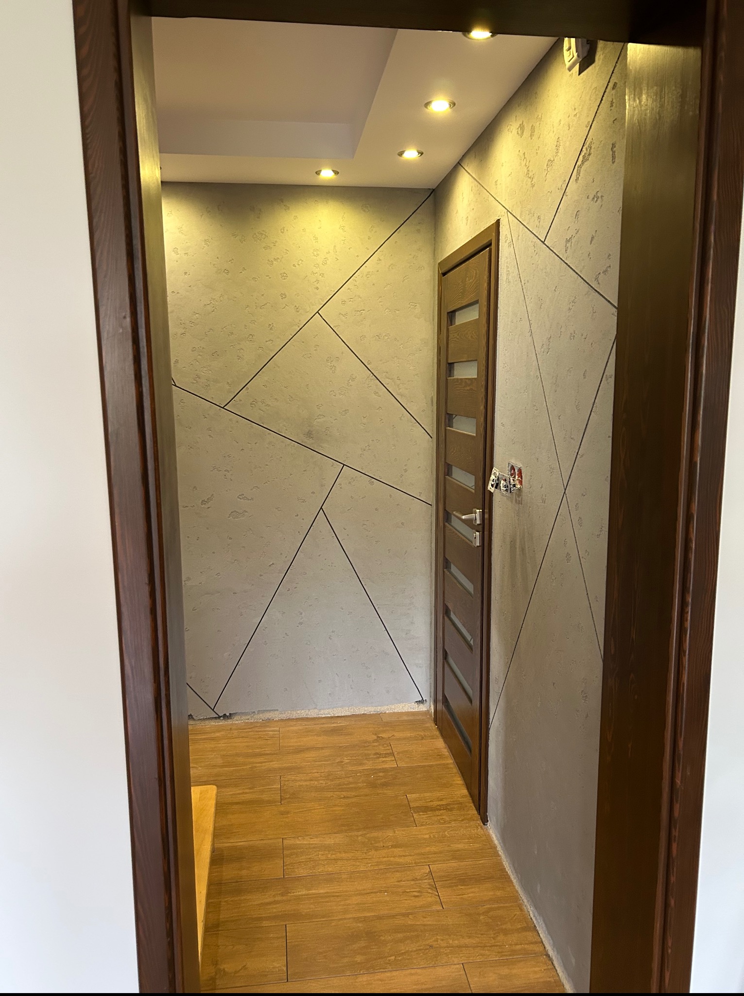 Wykończone wnętrze: korytarz z drewnianą podłogą, dekoracyjnymi ścianami w geometryczne wzory i nowymi drzwiami. Widok zza framugi.