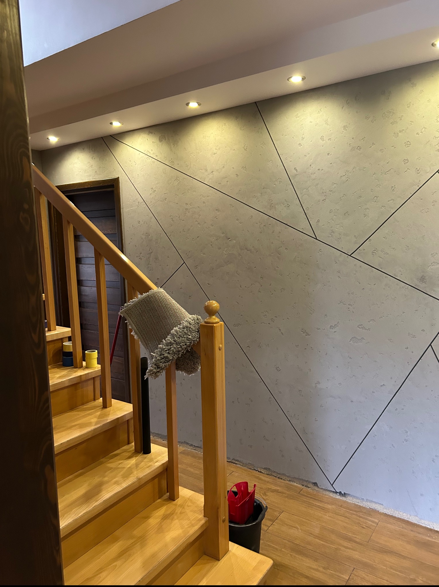 Wykończona ściana z geometrycznym wzorem i drewniane schody, oświetlone punktowo. W tle widoczne drzwi. Na schodach leżą narzędzia malarskie.