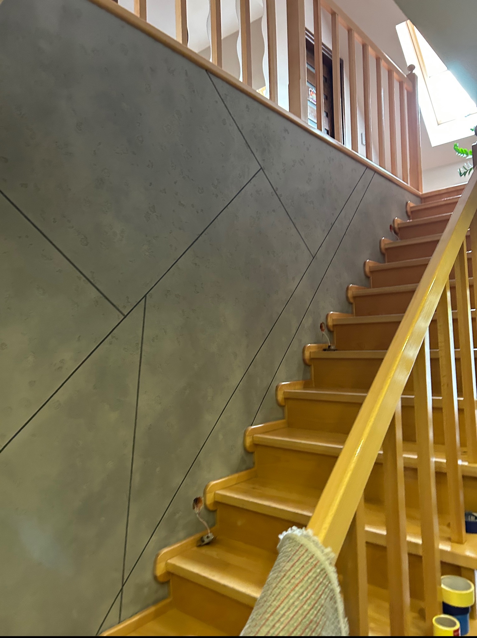 Ściana wykończona w geometryczny wzór obok drewnianych schodów z jasną balustradą. Widoczne elementy dekoracyjne i wykończeniowe na ścianie.