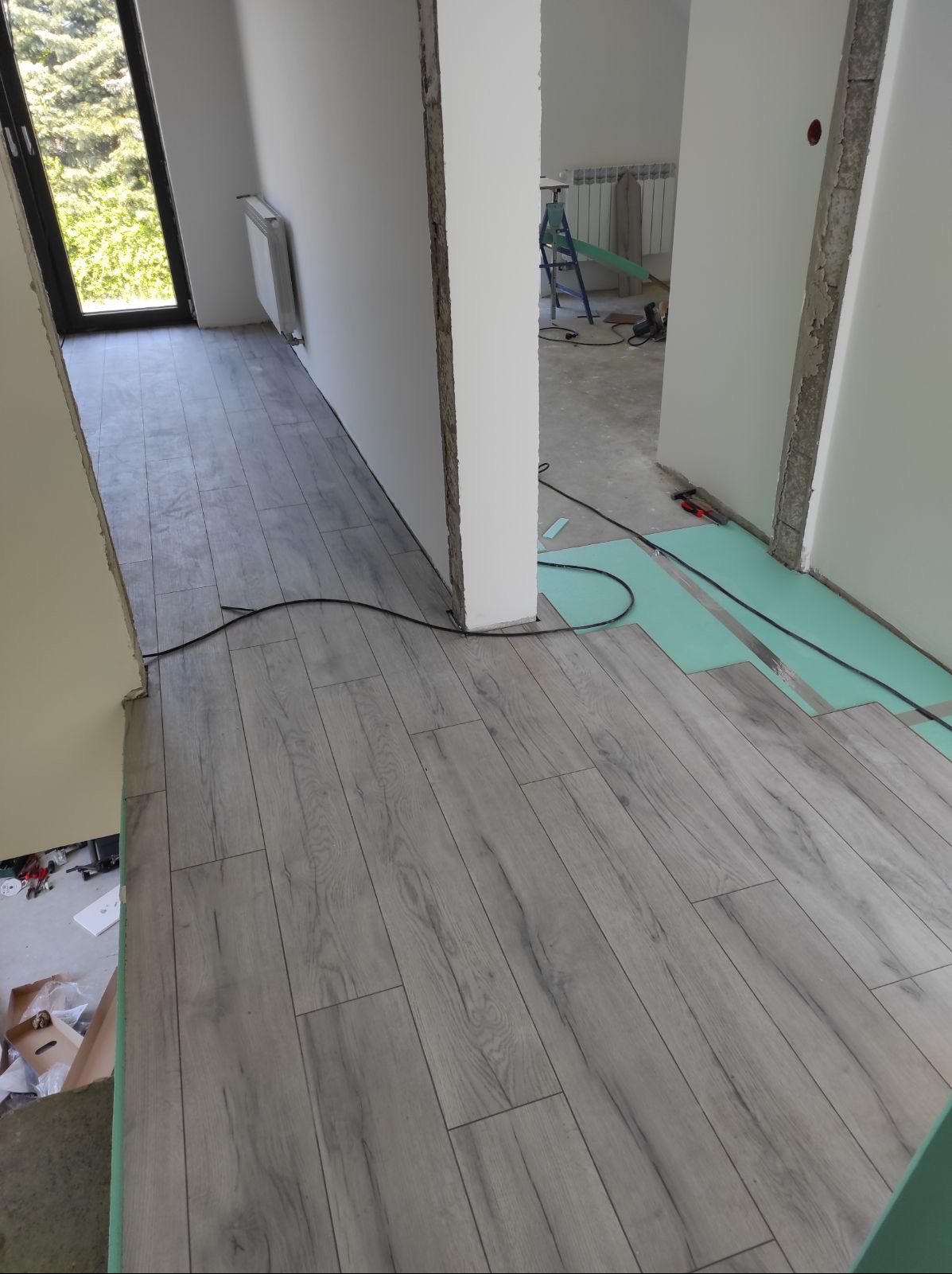 Układanie paneli podłogowych w jasnym odcieniu szarości, widoczne podłoże izolacyjne i narzędzia w tle. Częściowo wykończone pomieszczenie.