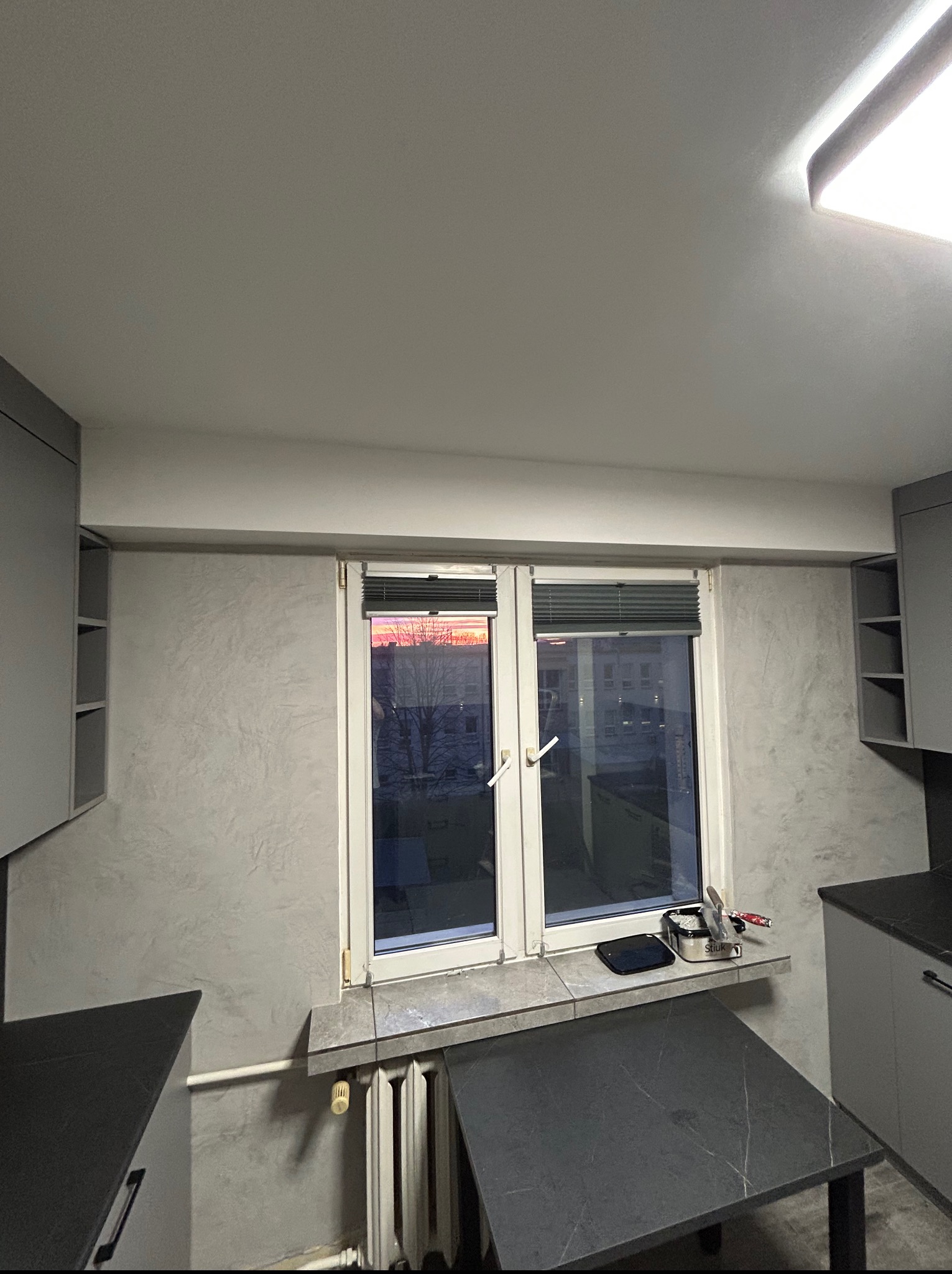 Szara kuchnia z oknem, widok na zachód słońca. Na parapecie akcesoria kuchenne. Ściana z efektem dekoracyjnym, minimalistyczne szafki.