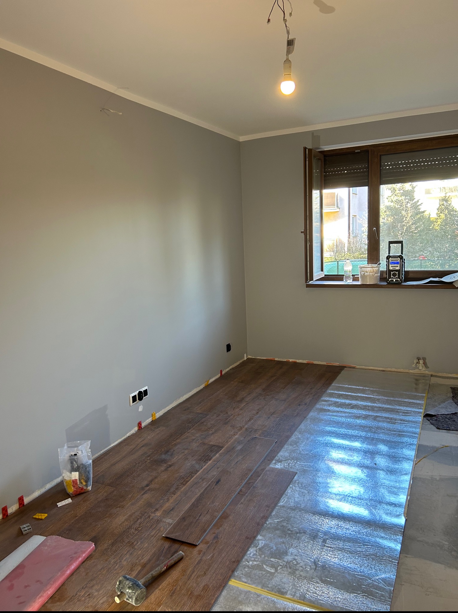 Wnętrze pokoju w trakcie układania paneli podłogowych. Część podłogi wyłożona folią izolacyjną, widoczne narzędzia: młotek, kliny dystansowe.