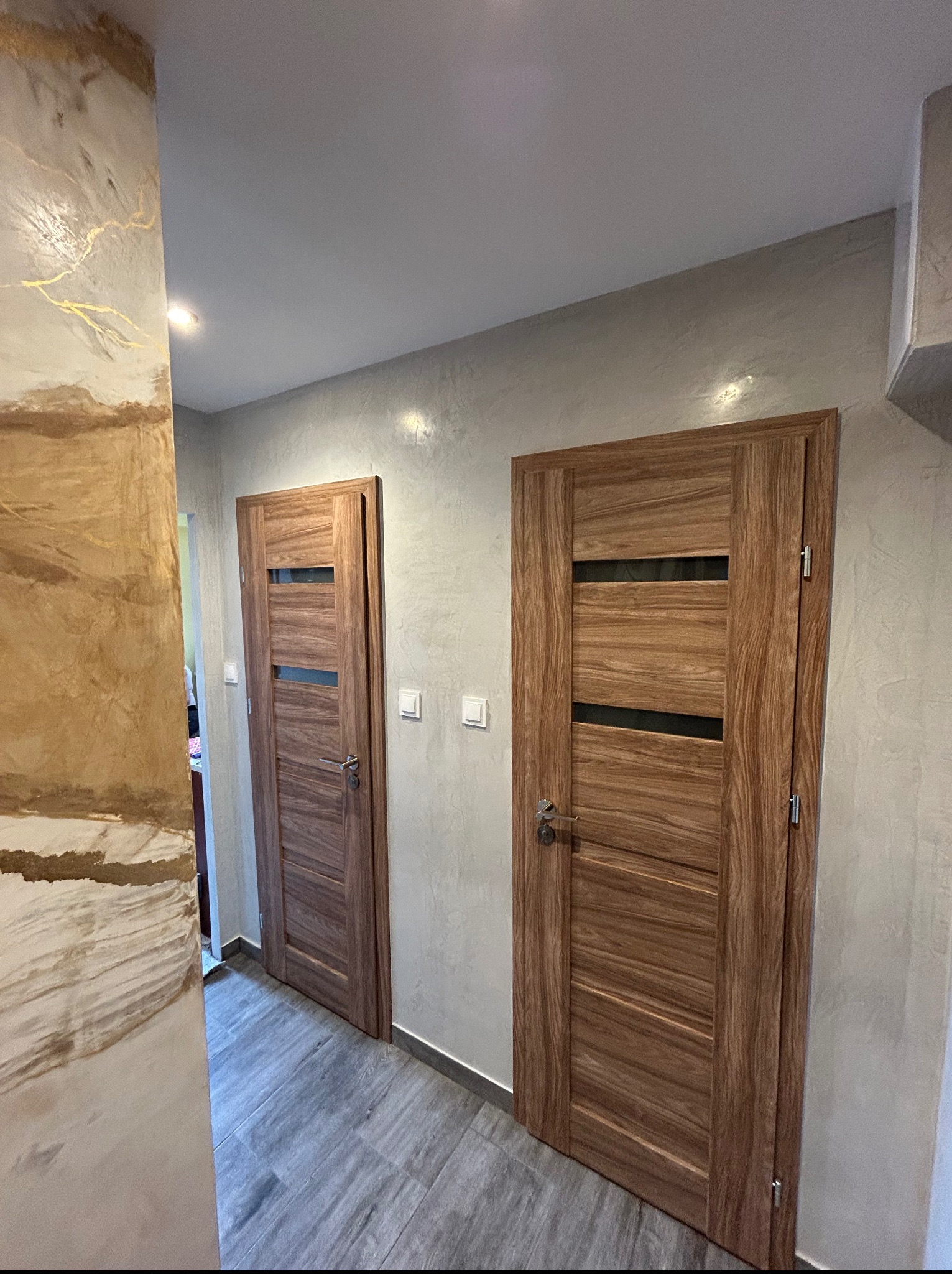 Dwa drewniane drzwi z przeszkleniami w szarej tonacji, na ścianie dekoracyjna faktura w odcieniach beżu i złota. Podłoga wyłożona szarymi panelami.