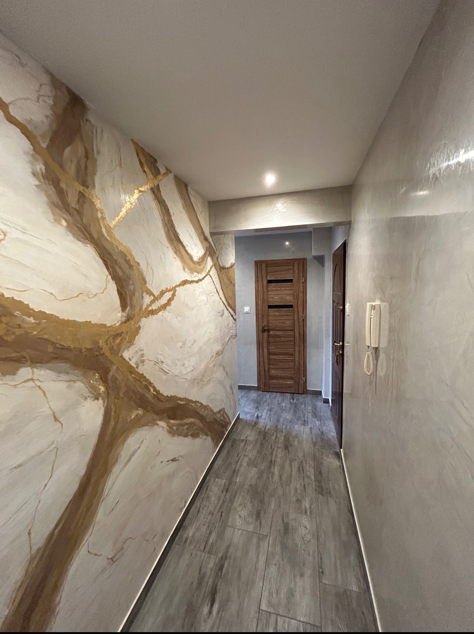 Korytarz po remoncie: ściana z efektem marmuru w odcieniach bieli i złota, szara podłoga imitująca drewno, drzwi z elementami drewna w głębi.