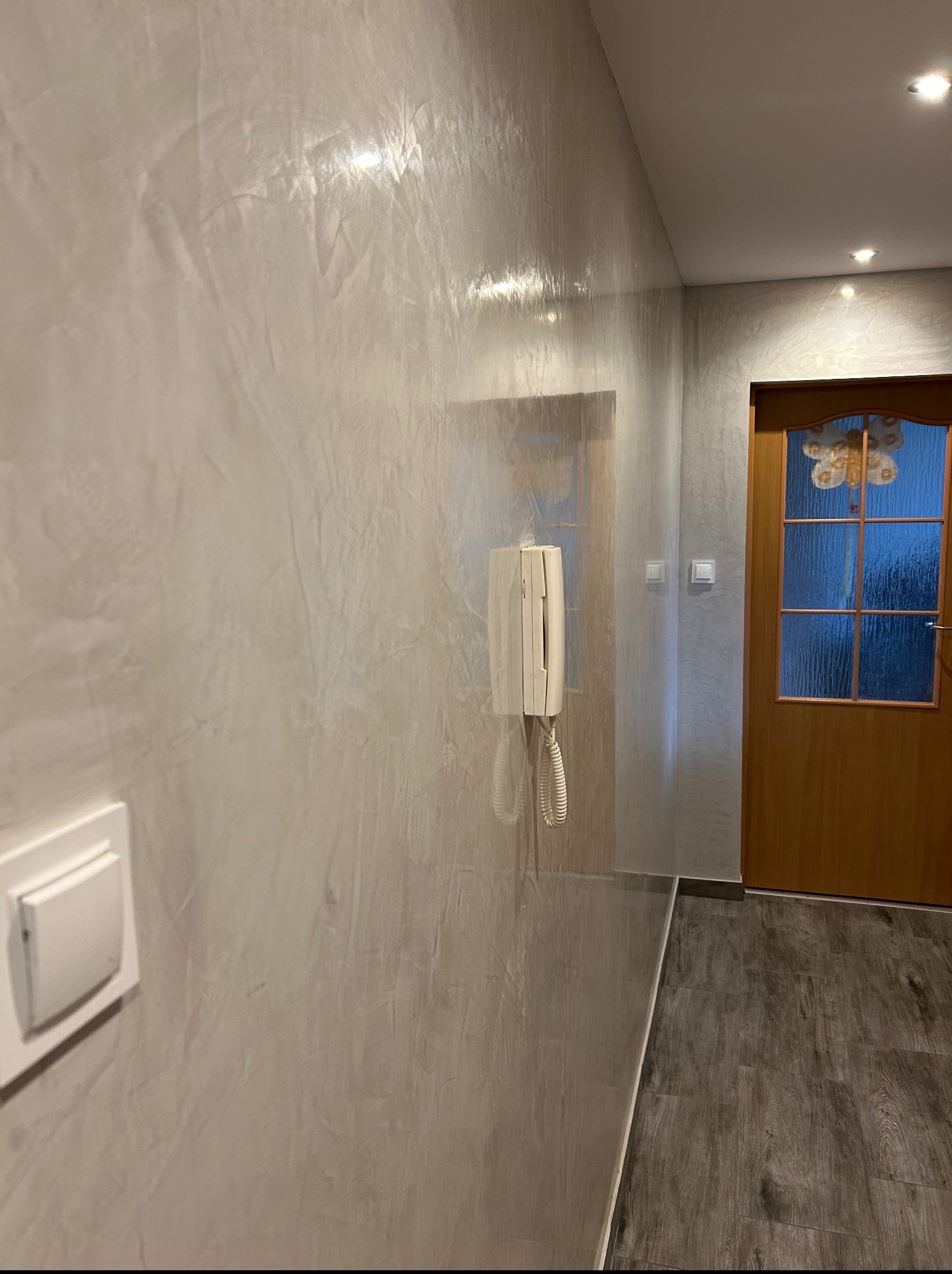 Korytarz po malowaniu z efektem dekoracyjnym na ścianie, widoczny domofon i drewniane drzwi z szybą. Podłoga wyłożona panelami.