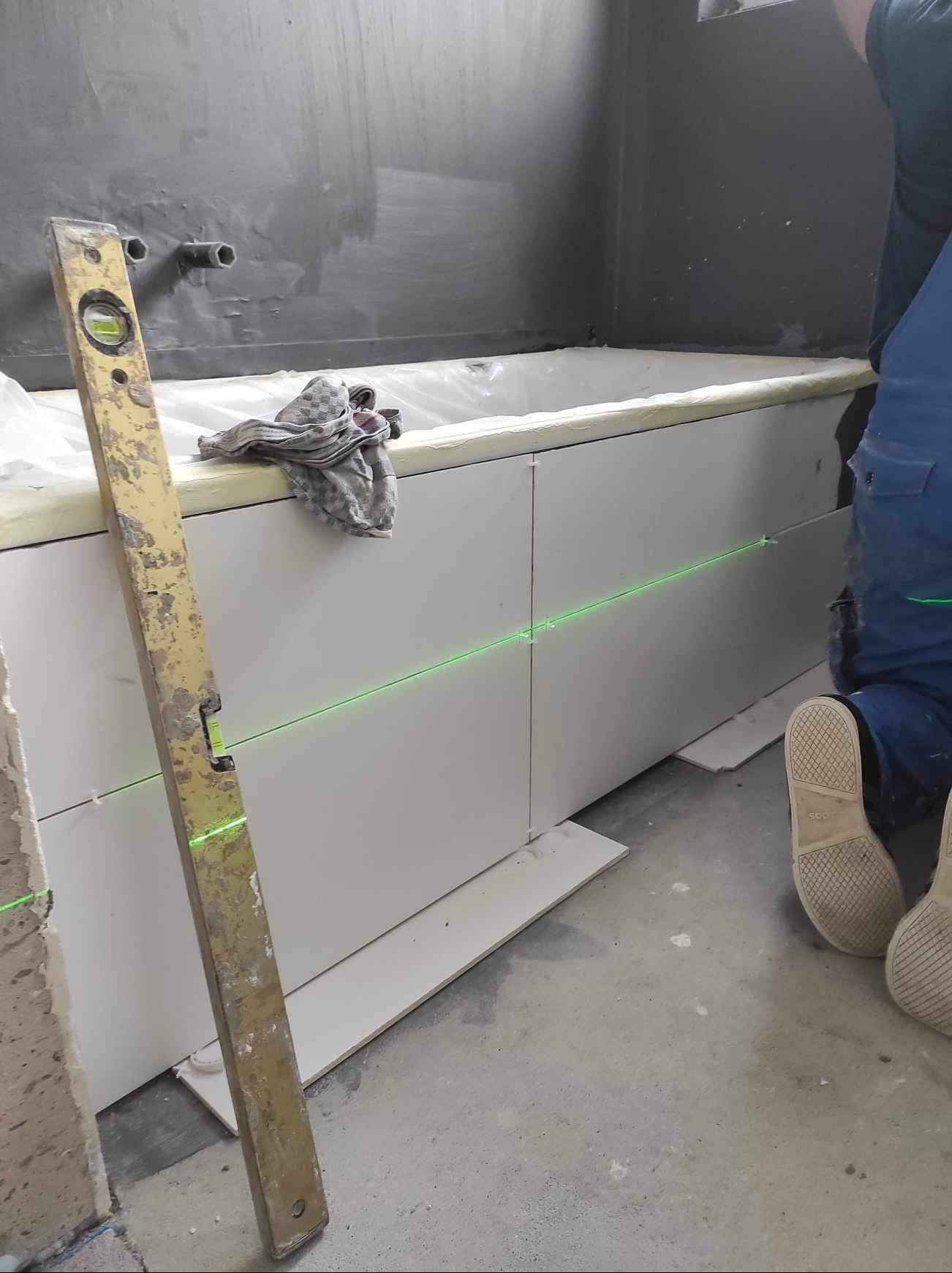 Układanie białych płytek na obudowie wanny, poziomica pionowa, linia lasera wyznacza poziom. Fragment osoby w niebieskim kombinezonie glazurnika.