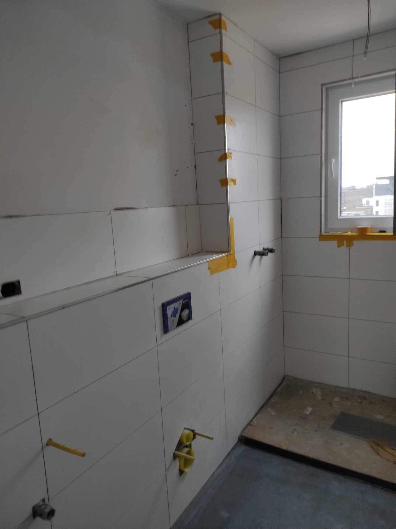 Białe płytki w łazience w trakcie montażu, zabezpieczone żółtą taśmą. Widoczne wystające rury i puszka pod przycisk spłuczki. Okno w tle.