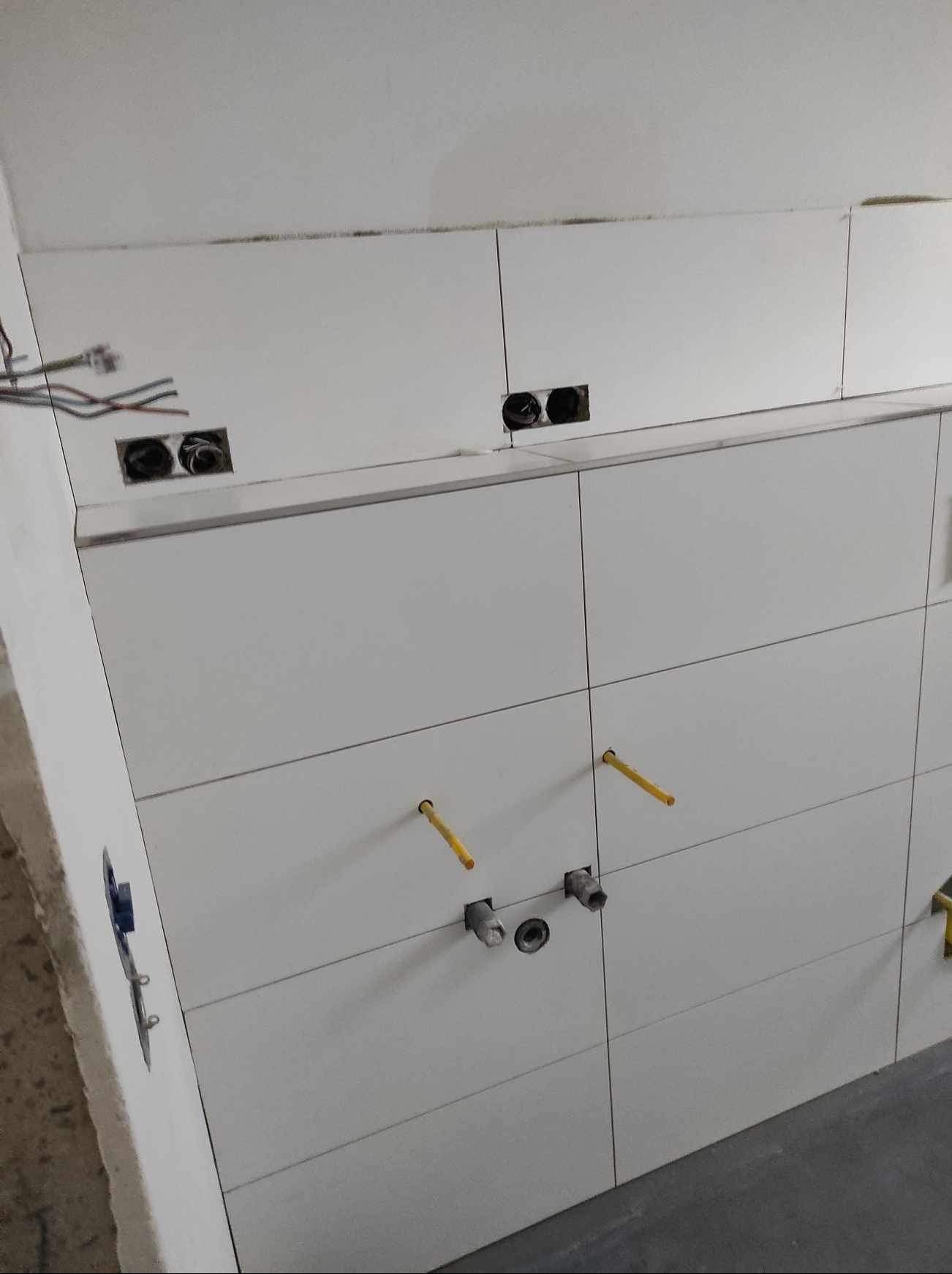 Białe, poziome płytki na ścianie z widocznymi otworami na gniazdka i rury, w trakcie prac wykończeniowych. Widoczne przewody elektryczne i fragment posadzki.