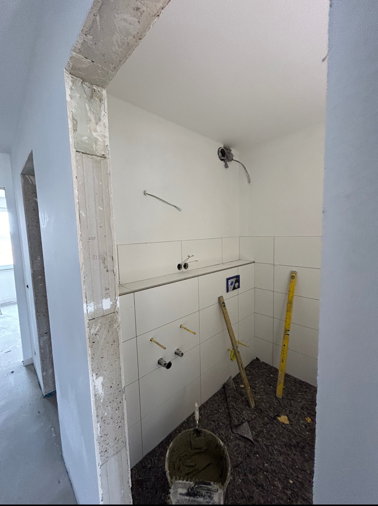 Wnętrze łazienki w trakcie prac wykończeniowych: białe płytki ułożone do połowy ściany, widoczne rury, poziomice, wiadro z zaprawą. Surowy betonowy portal drzwiowy.