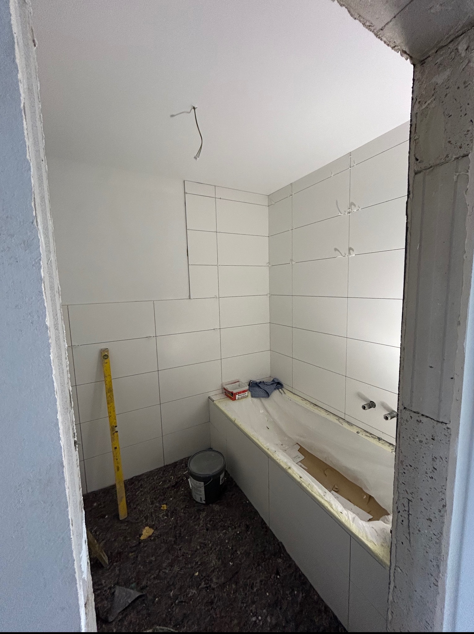 Biała łazienka w trakcie remontu: świeżo położone płytki na ścianach, przygotowana wanna, poziomica oparta o ścianę, widoczne kable z sufitu. Surowy stan wykończenia.