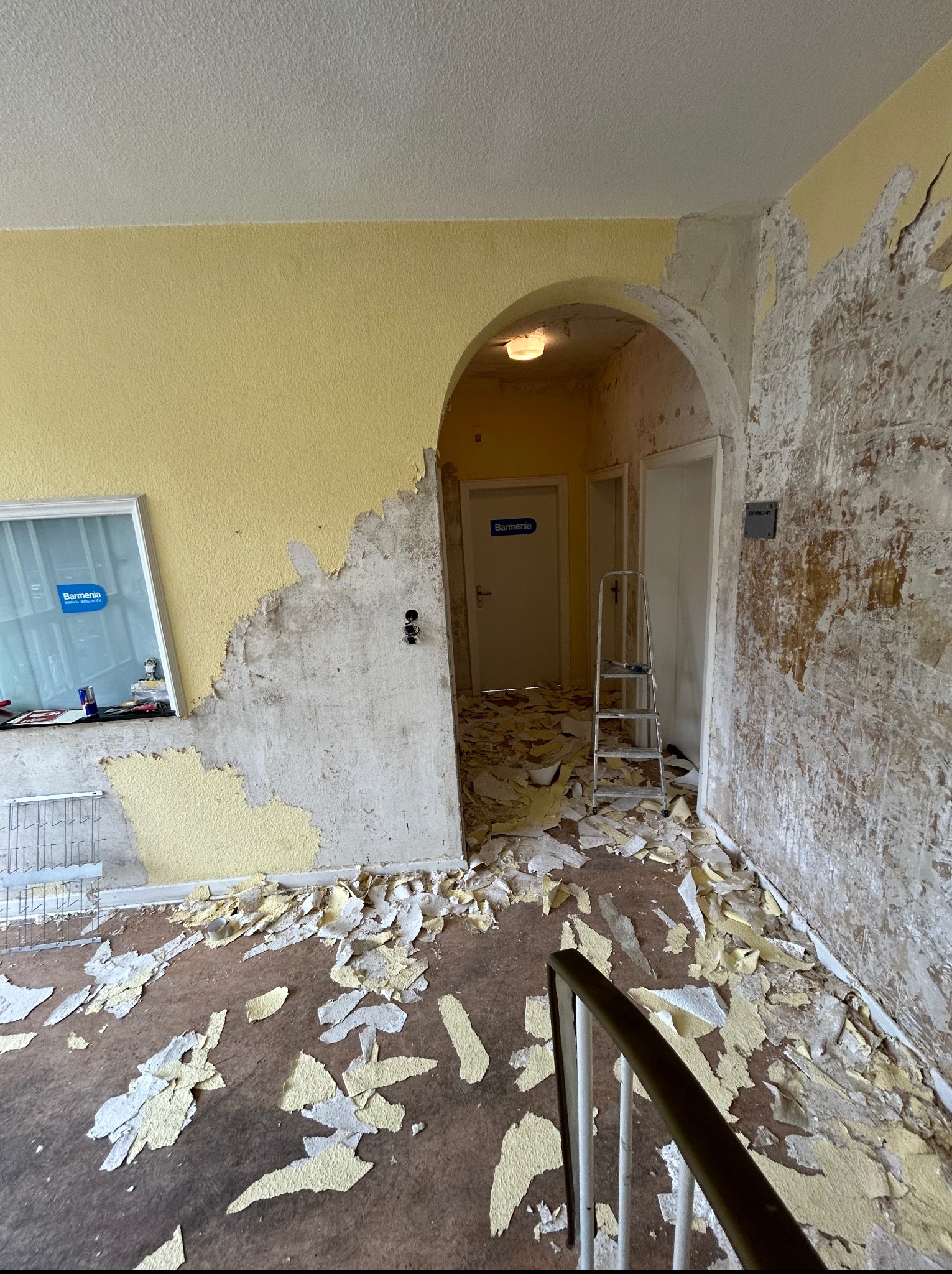 Pokój w trakcie usuwania starej farby, widoczne fragmenty żółtej ściany, resztki farby na podłodze i drabina w przejściu z łukiem. Przygotowanie do malowania.