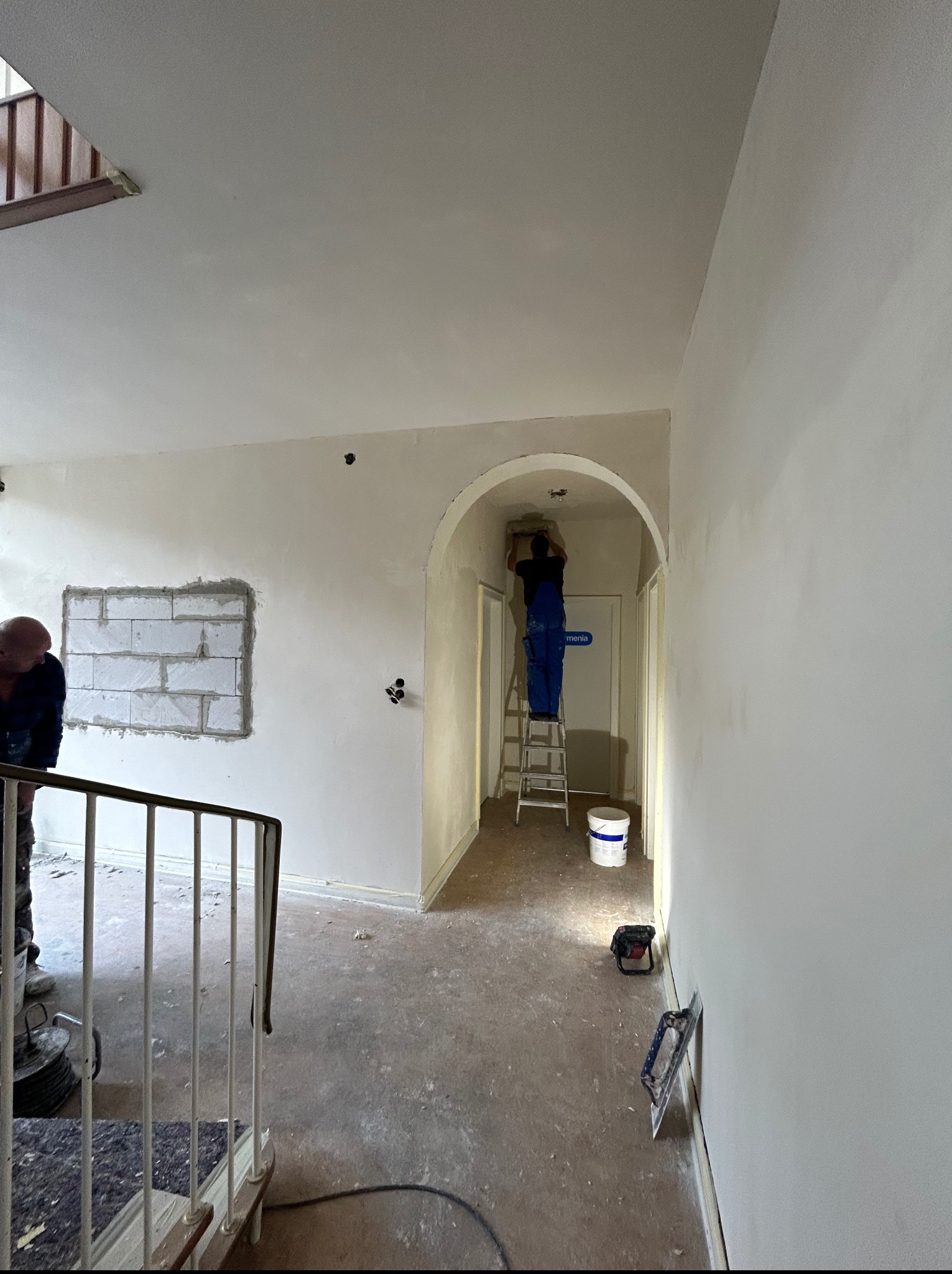 Wnętrze w trakcie prac wykończeniowych: szpachlowanie ścian przy użyciu drabiny, widoczny fragment ściany z bloków i narzędzia pracy. Jasne, naturalne światło.