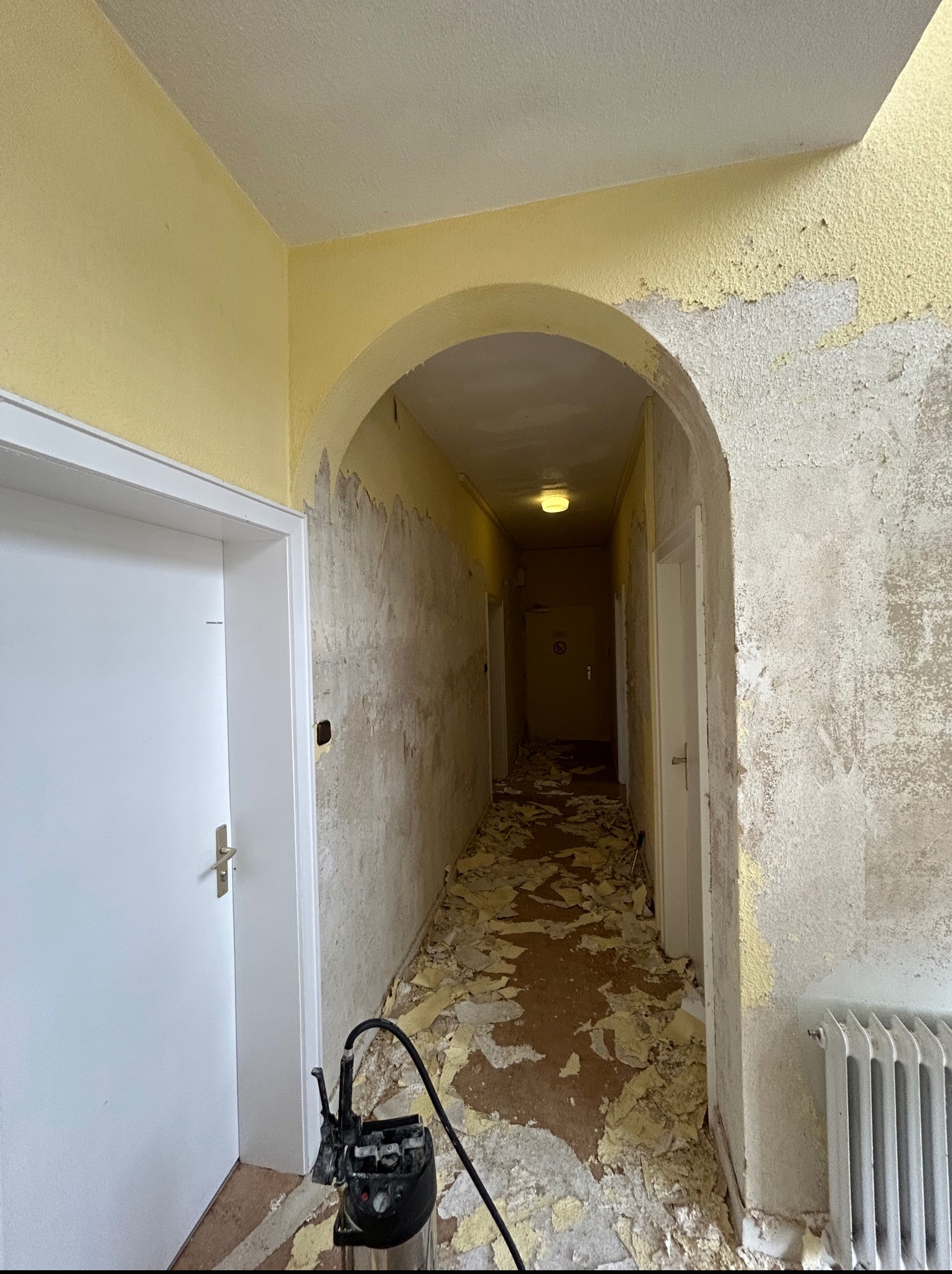 Korytarz w trakcie usuwania starej farby ze ścian i sufitu, widoczne resztki farby na podłodze, narzędzie do usuwania farby przy drzwiach, grzejnik i łukowe przejście.