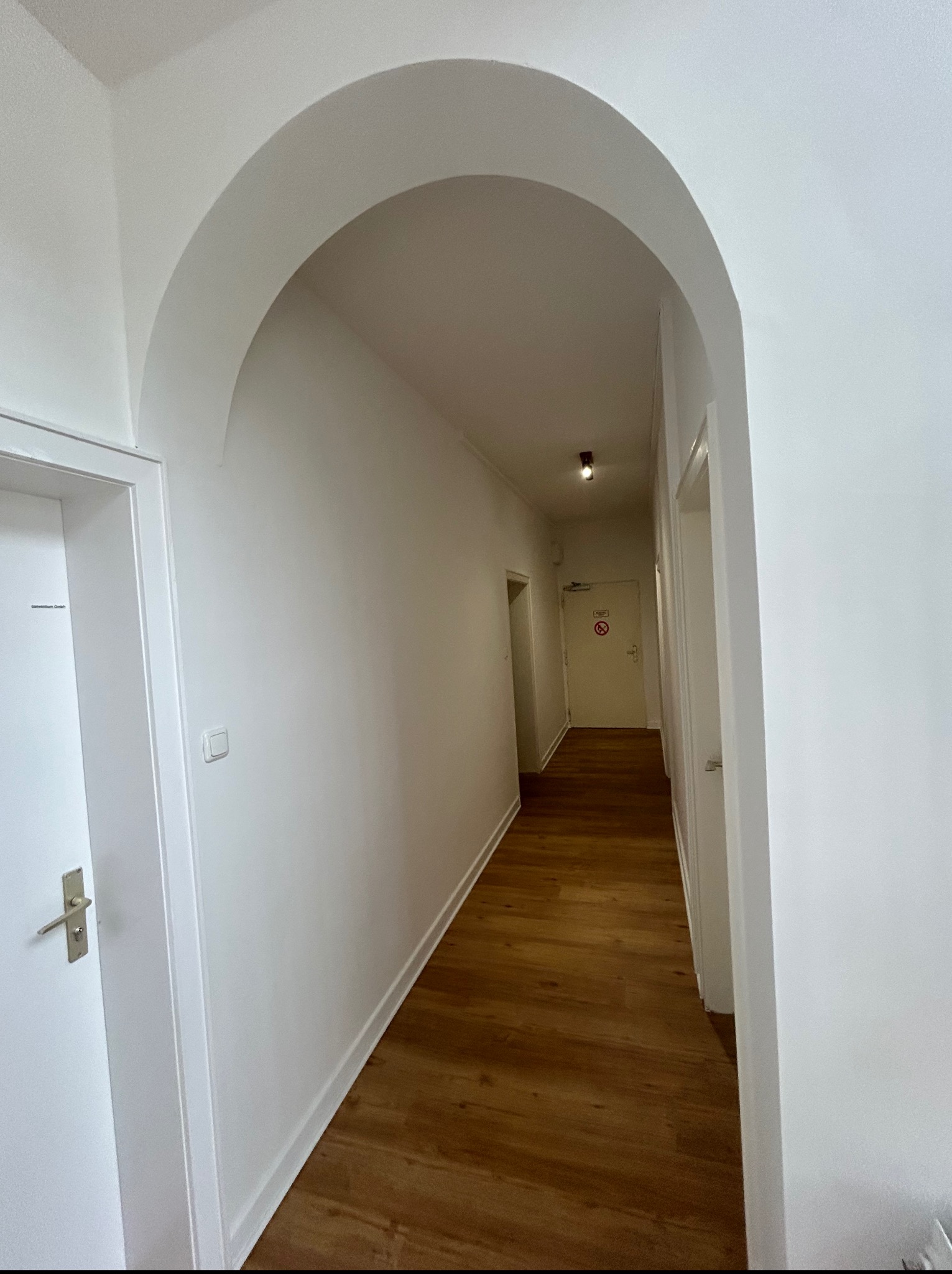 Biały korytarz z łukowym przejściem i drewnianą podłogą. Widok na drzwi na końcu korytarza. Minimalistyczny design, świeżo po remoncie.