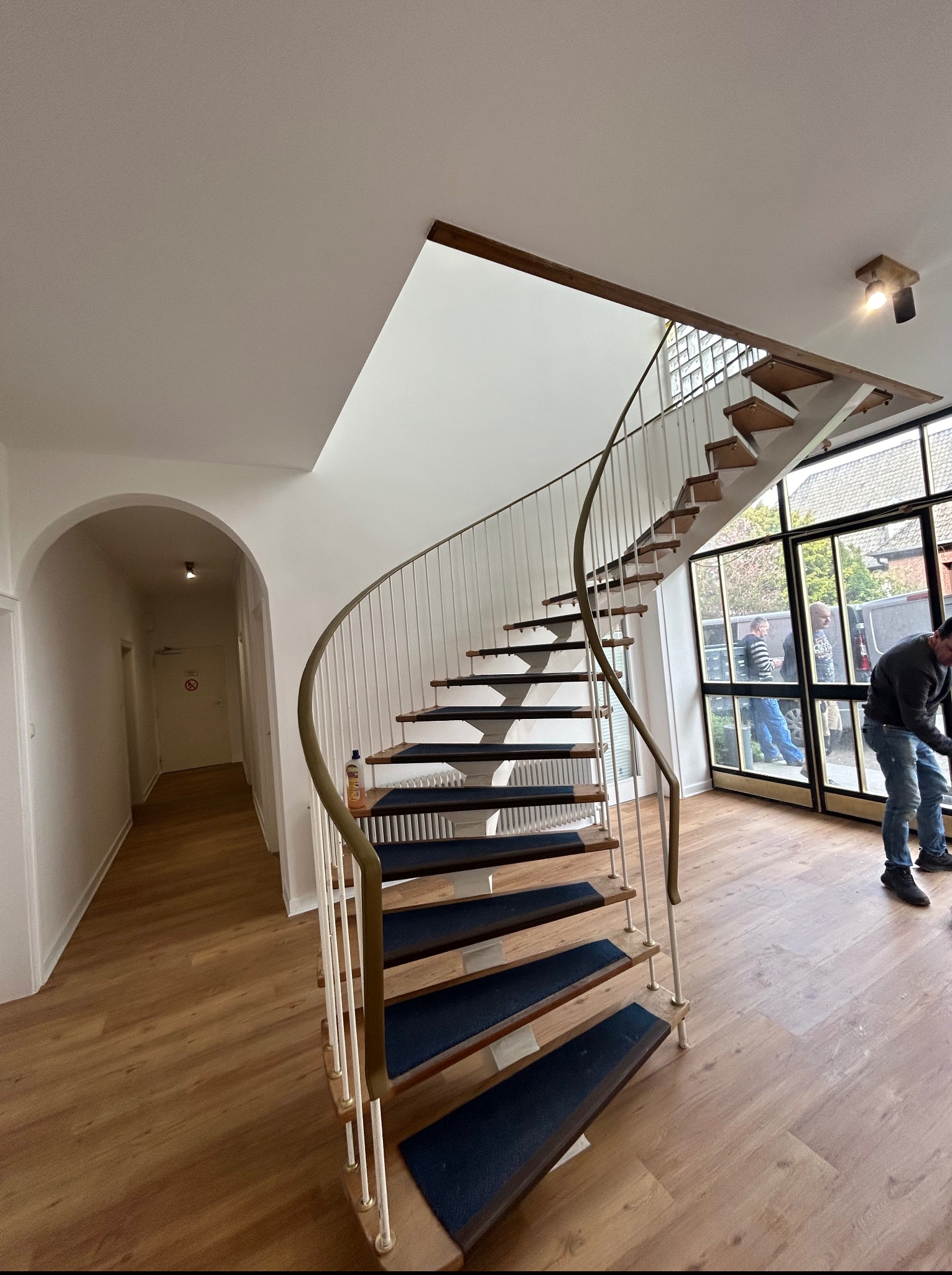 Spiralne schody z drewnianymi stopniami i białymi balustradami w jasnym wnętrzu. Widoczny fragment podłogi i okna z widokiem na zewnątrz. Osoba w tle.